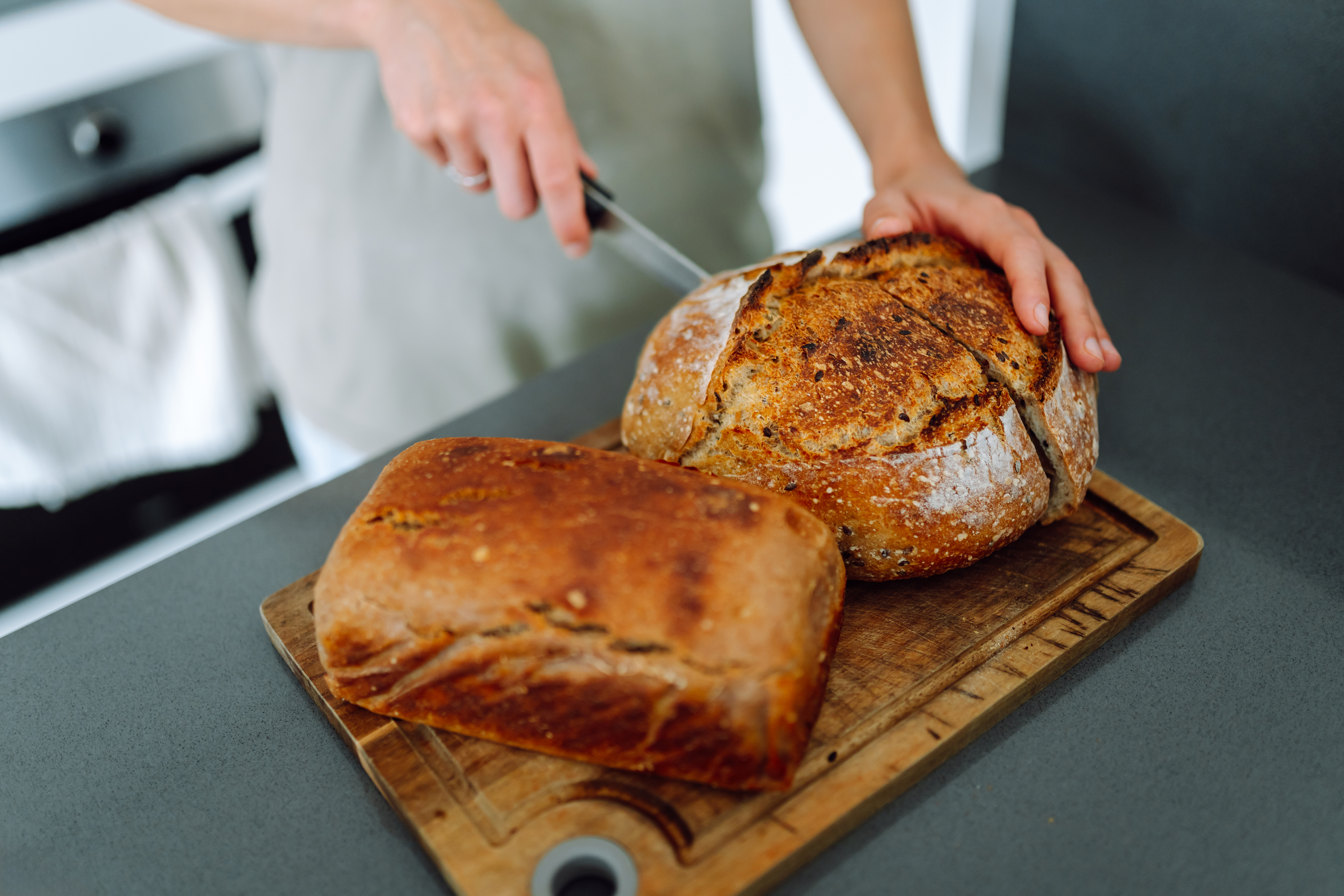 Close-up of a young female baker slicing homemade sourdough bread on a wooden board. A housewife stands in a modern kitchen near freshly baked bread. The concept is bakery, indoors, and cozy.